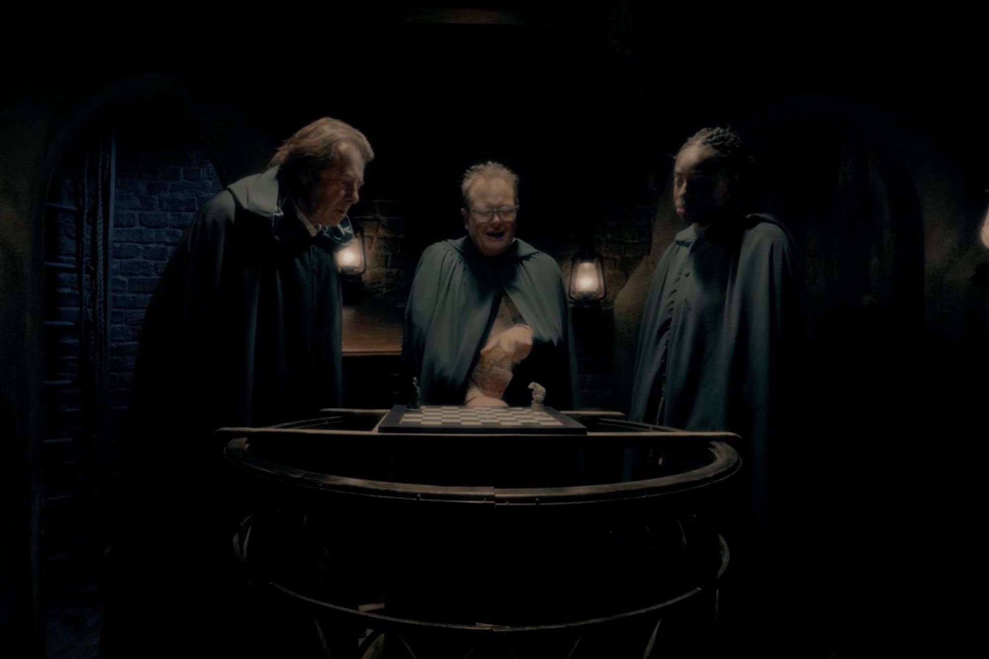An image collage containing 1 images, Image 1 shows Three people in cloaks looking at a chessboard in a dimly lit room