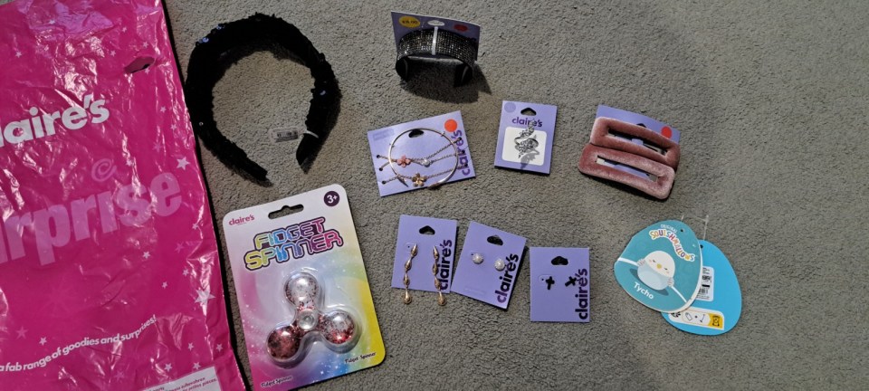 A collection of Claire's accessories and toys on a gray carpet, including a pink shopping bag, a black sequin headband, a rhinestone bracelet, two necklaces, a fidget spinner, three pairs of earrings, a snake ring, two velvet hair clips, and a Squishmallows tag for Tycho.