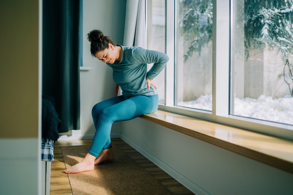 Woman suffering from back pain while sitting near a window with a snowy view.