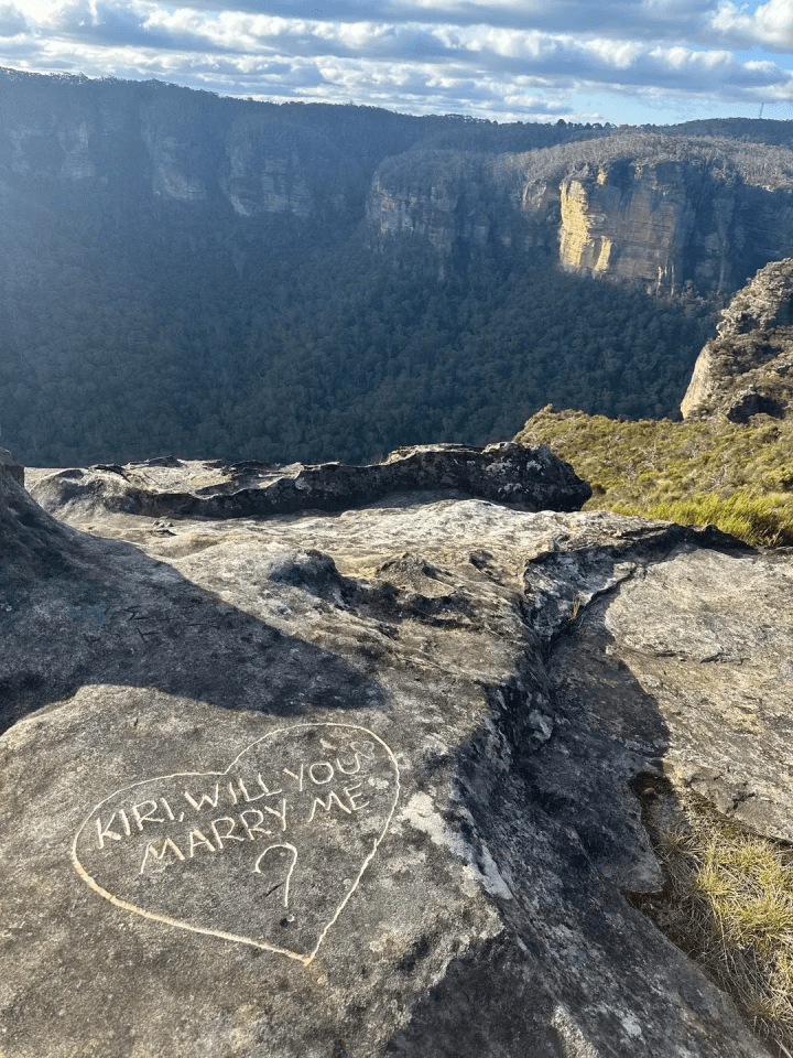 "Kiri, will you marry me?" carved into a heart shape on a rock overlooking a forested valley and cliff face.