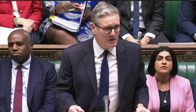 Keir Starmer speaking at a podium with two people sitting beside him.