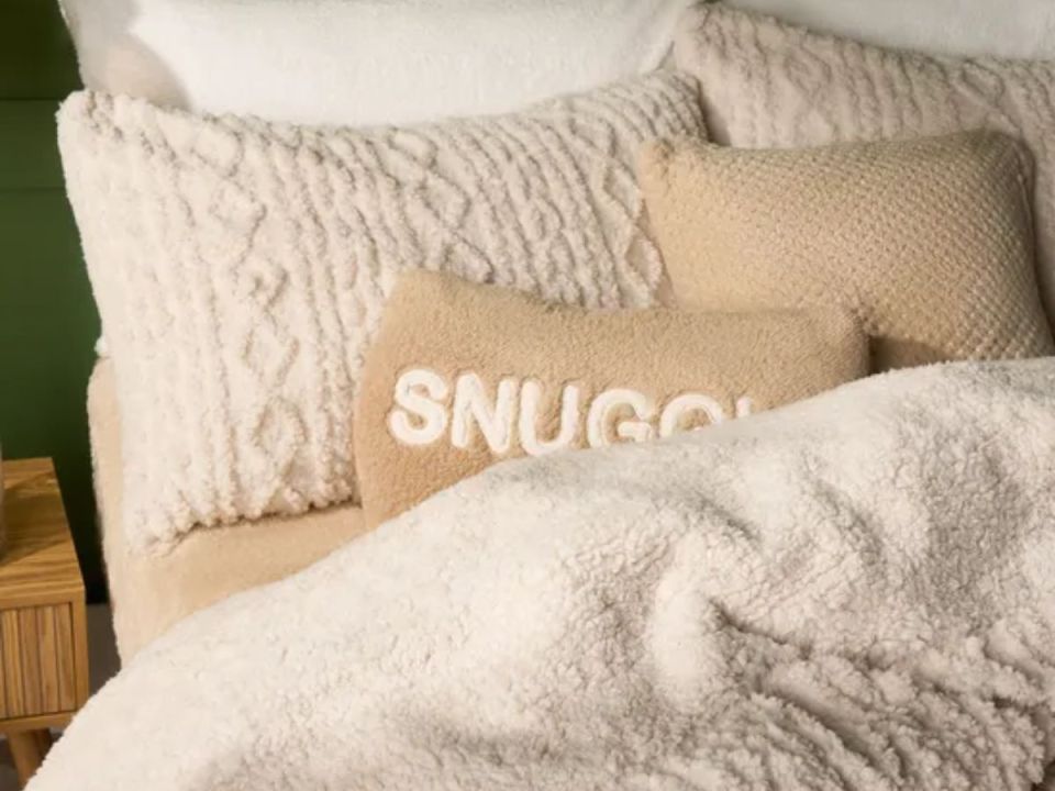 A bed with cream-colored pillows and a blanket, one pillow embroidered with the word "SNUGGLE".