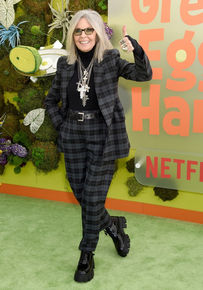 Diane Keaton smiling, wearing a black turtleneck and plaid pantsuit, giving a thumbs-up on a green carpet with "Green Eggs and Ham" and Netflix logos in the background.