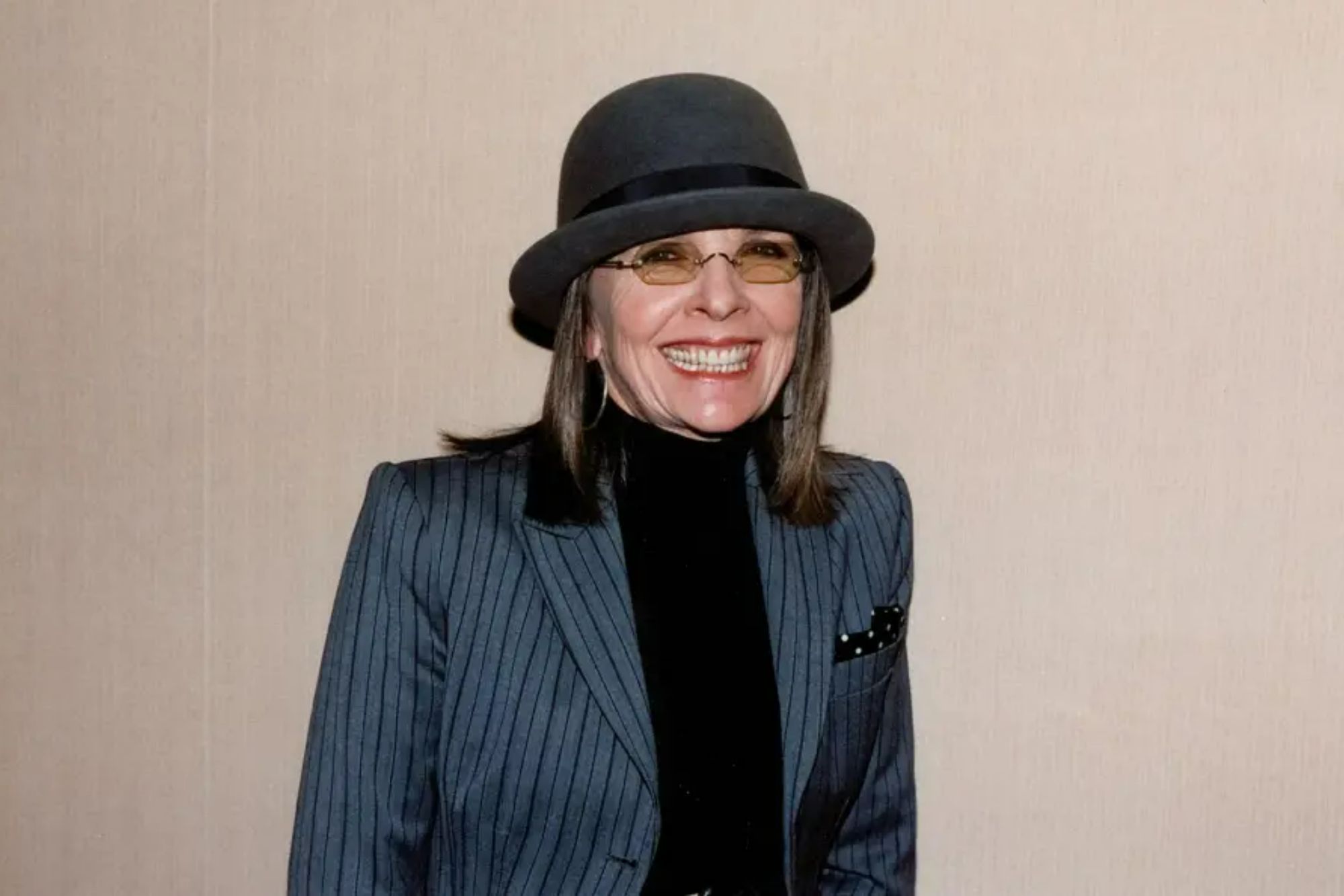 An image collage containing 1 images, Image 1 shows Diane Keaton wearing a pinstripe blazer, black turtleneck, a dark gray fedora, and amber-tinted glasses