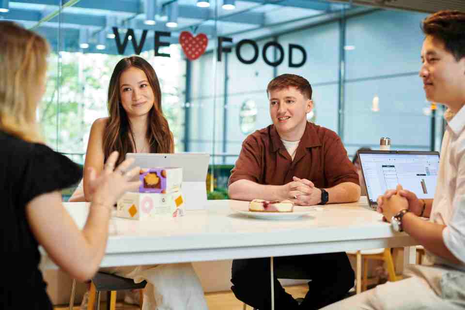 Four people sitting at a table with pastries and a laptop, in front of a "WE ❤️ FOOD" sign.