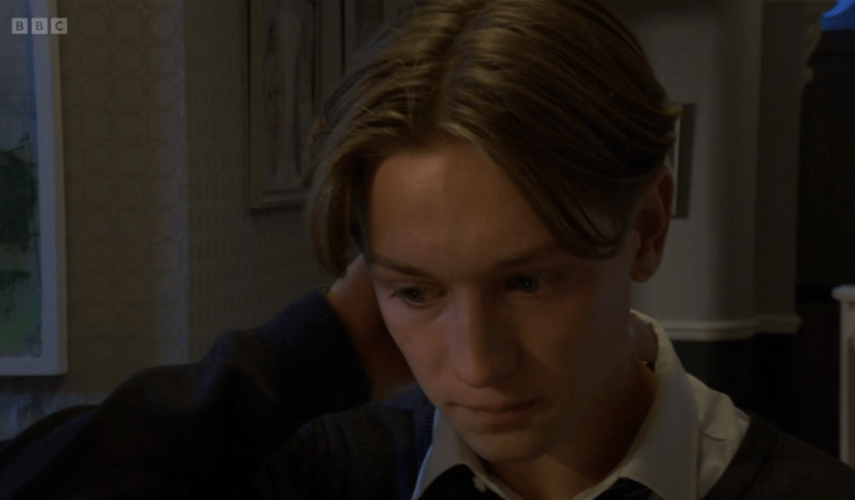 A young man with light brown hair looking down, with his hand touching his neck.