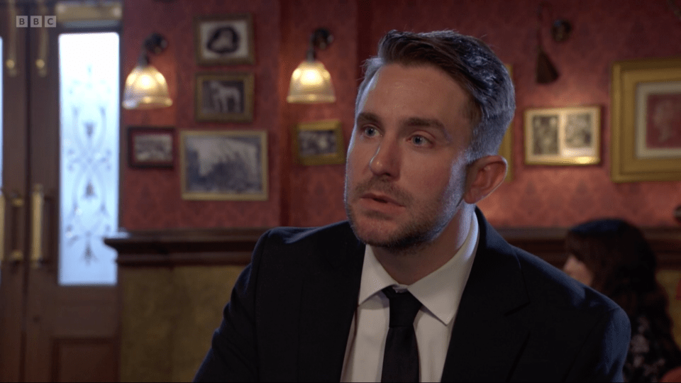A man in a black suit and tie, with a short beard, looks intently to the left.