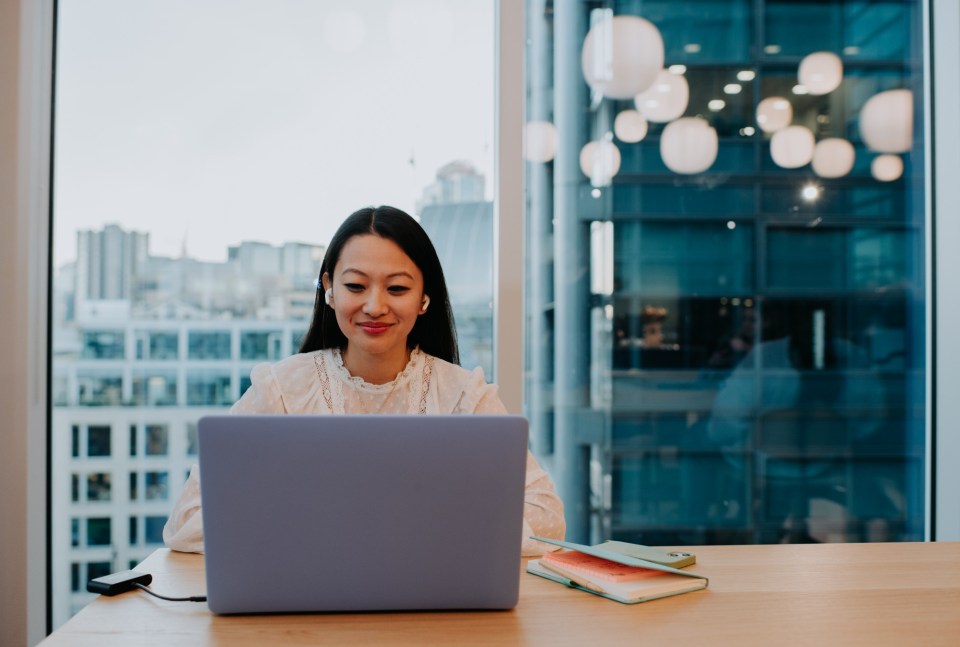 A young woman in a modern office space uses a laptop at a desk. Space for copy.