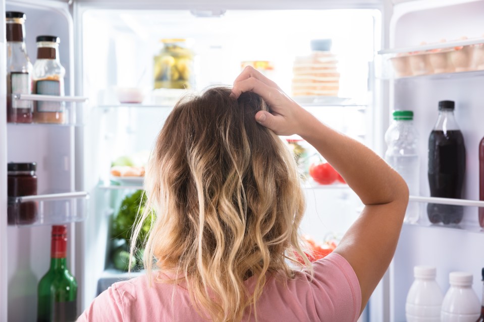 Rear view of a confused woman looking into an open refrigerator, scratching her head.
