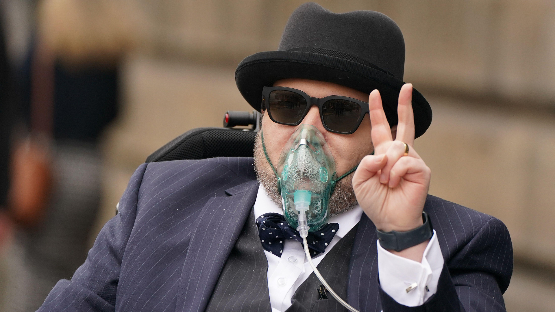 A man wearing a hat, sunglasses, an oxygen mask, and a pinstriped suit makes a peace sign.