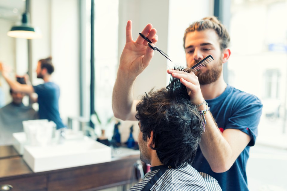 A man with a beard and hair in a bun giving a customer a haircut with scissors and a comb.