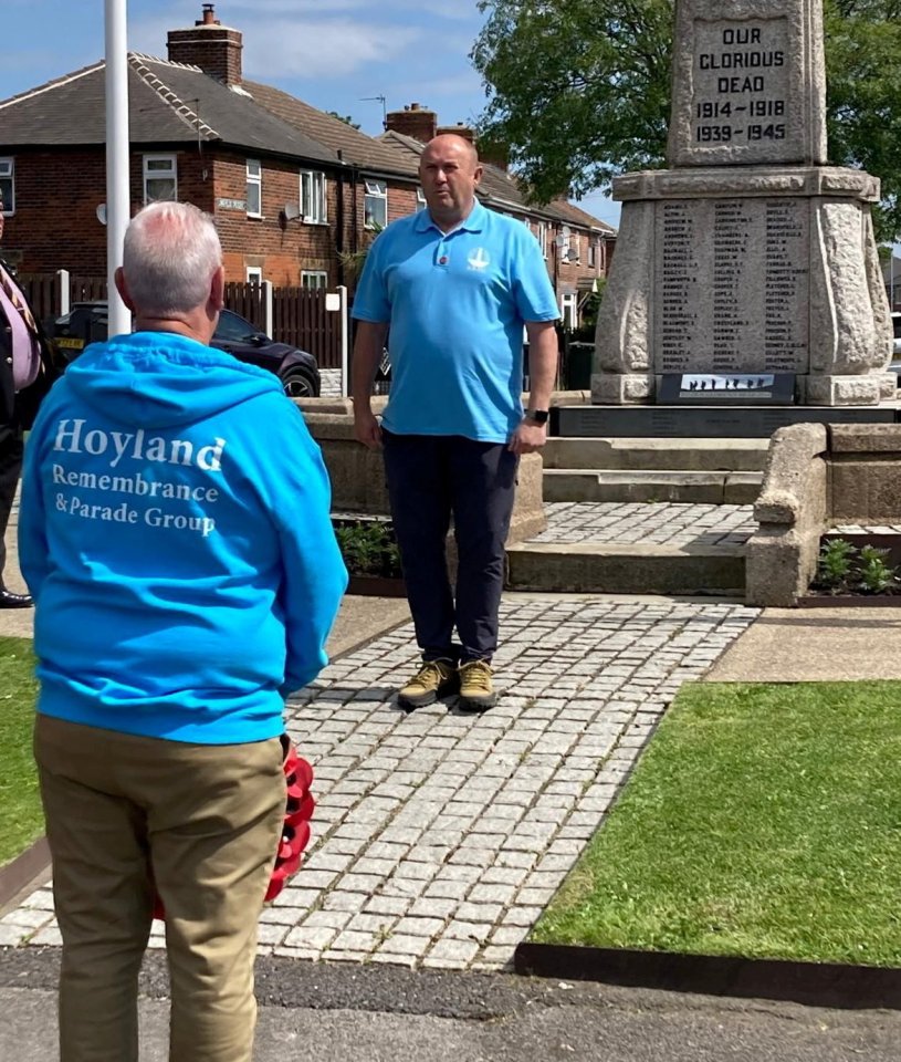 Two men stand near a war memorial, one wearing a blue hoodie that reads "Hoyland Remembrance & Parade Group."
