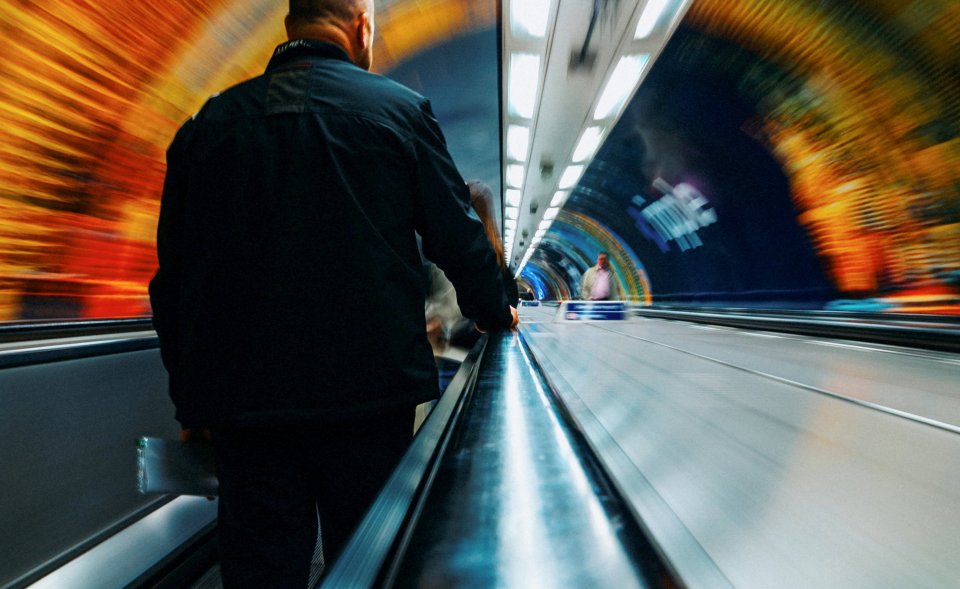 A person in a black jacket rides a moving walkway in a brightly lit tunnel.