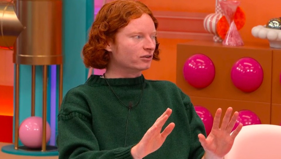 George with red curly hair, wearing a green sweater, holding his hands up, during Big Brother.