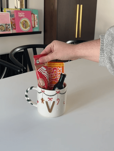 A hand placing a Maltesers hot chocolate packet into a festive white mug with a gold "V" and a miniature bottle of Baileys.