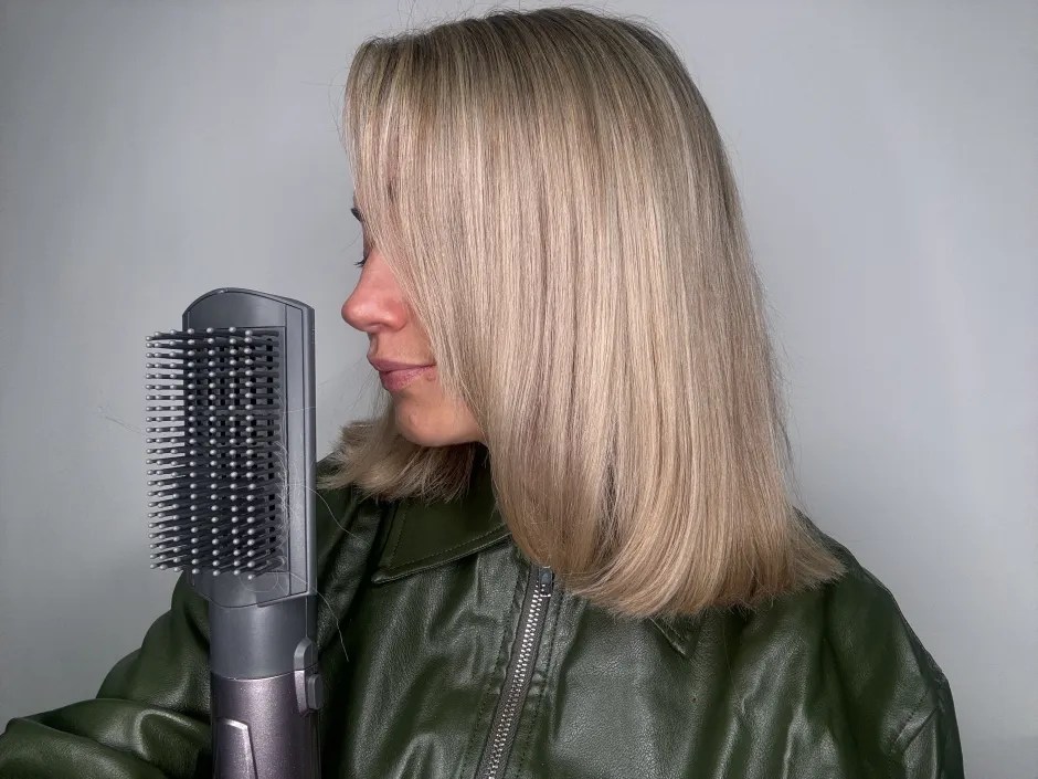 A woman with straight blonde hair holds a hairbrush with some loose strands of hair on its bristles.