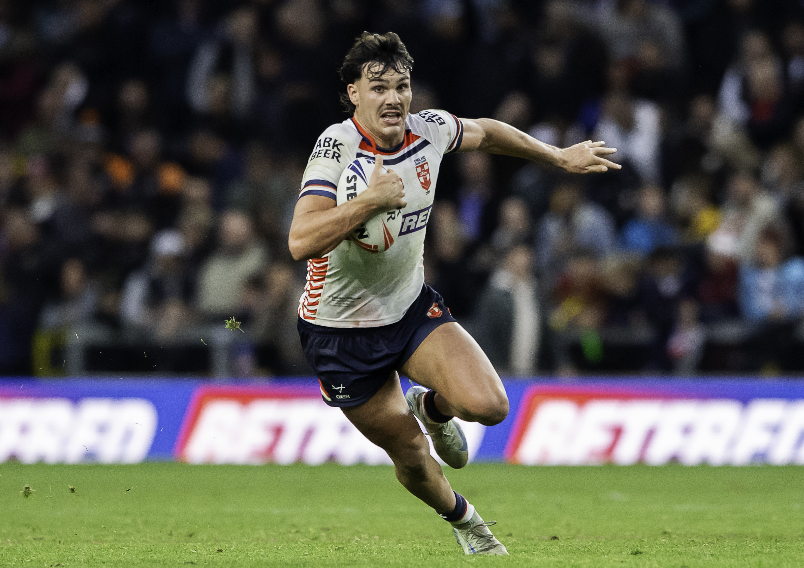 A rugby player in a white and red jersey with a rugby ball running on the field.