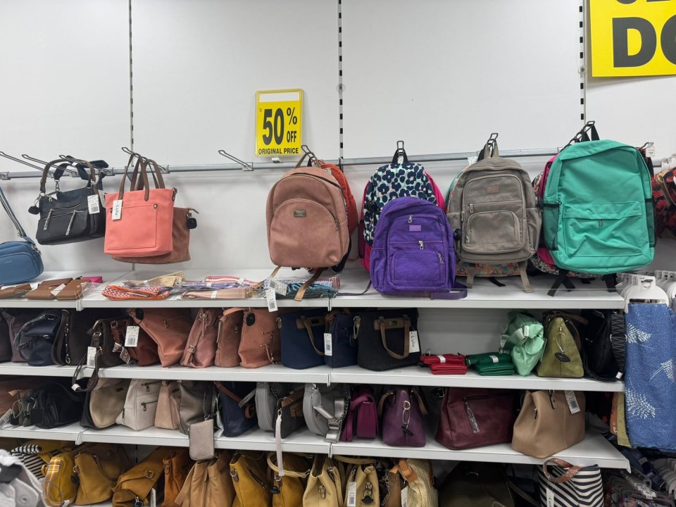 A store shelf filled with bags and backpacks of various colors and styles, with a "50% off original price" sign prominently displayed.