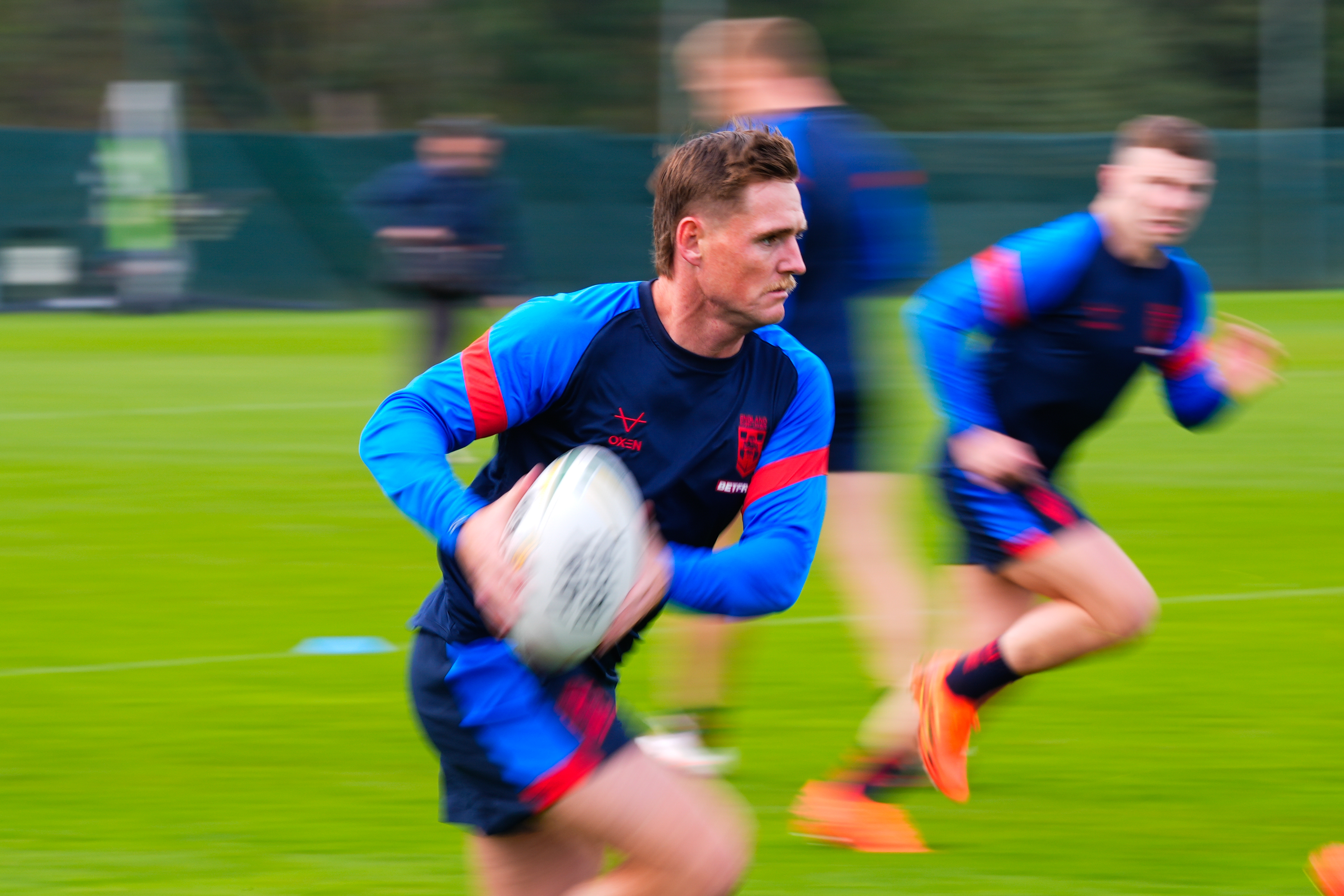 A rugby league player with a mustache runs with the ball on a grassy field, with other blurred players in the background.