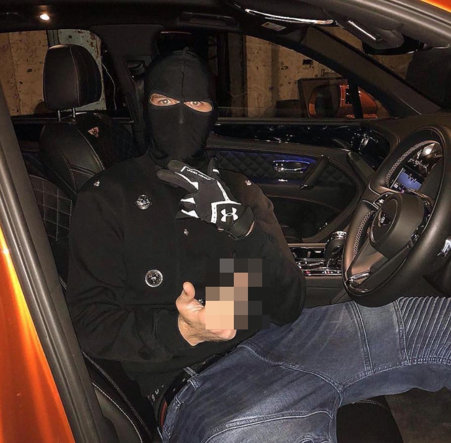 A person in a black balaclava, black jacket, and gloves, sits in the driver's seat of an orange car, holding a blurred object in one hand.