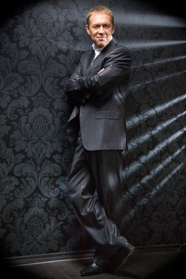 Andrey Lugovoy, in a suit, stands with arms crossed against a dark patterned wall.