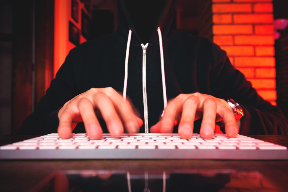 Person in a black hoodie with a covered face typing on a computer keyboard.