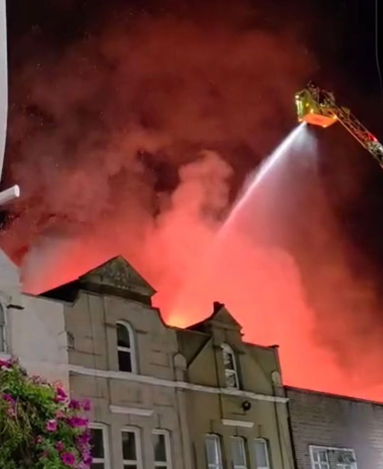 A fire engine with an extended ladder directs a stream of water onto a burning building at night, with flames and smoke visible.