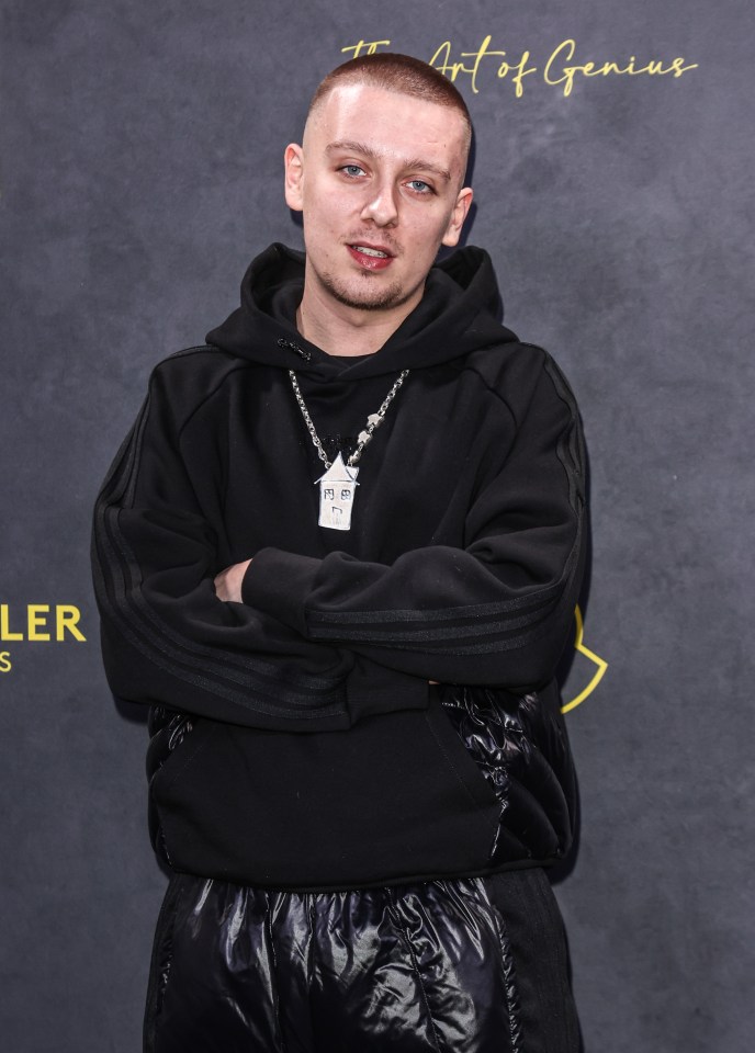 Aitch at the Moncler Genius show in a black hoodie and shiny black pants, with a house-shaped pendant.