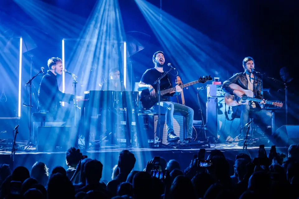 Kodaline performing live on stage, with Steve Garrigan at a piano, Jason Boland playing bass, and Mark Prendergast playing acoustic guitar, all under blue stage lights.