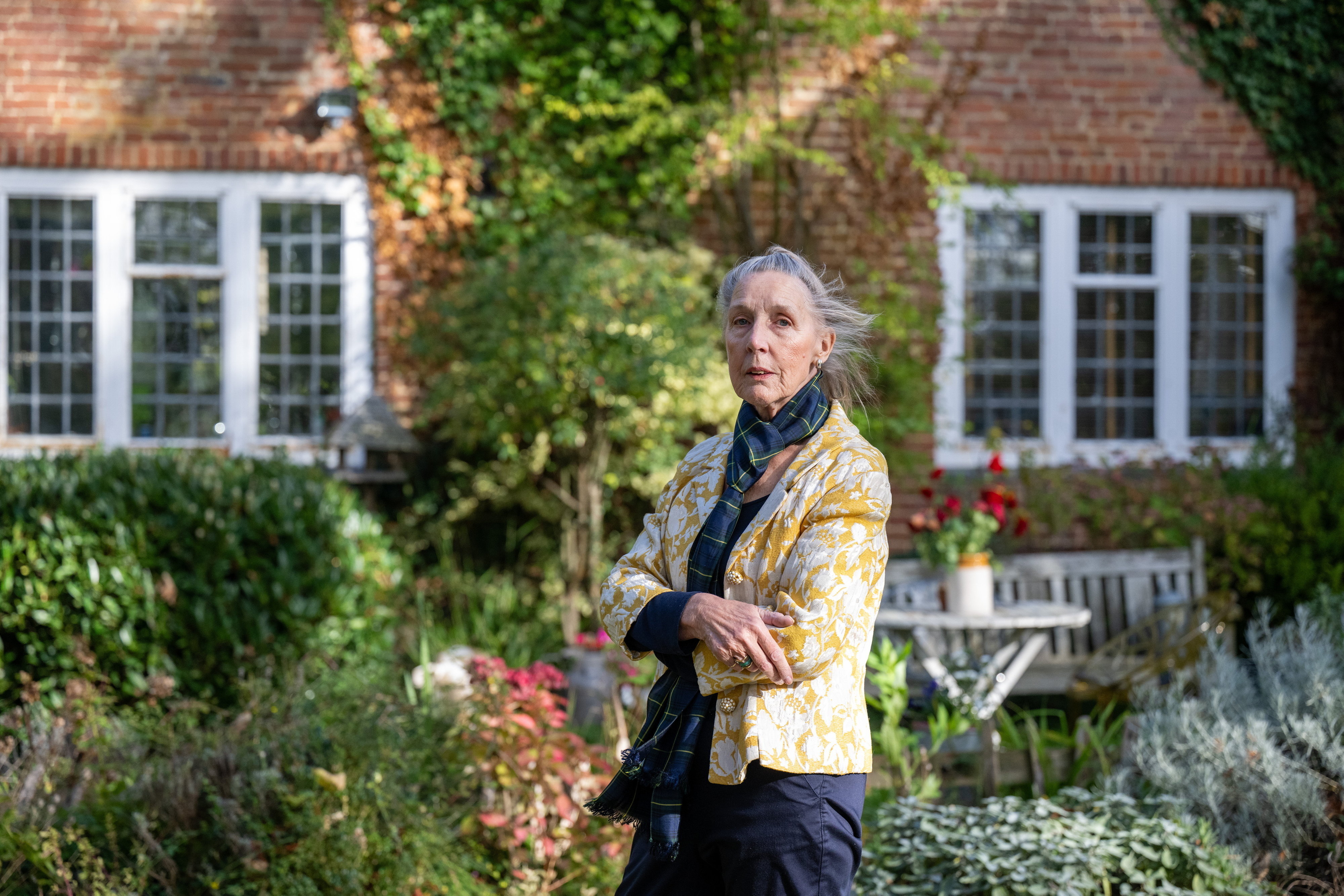 Baschea Walsh, 71, stands in front of a brick cottage with windows and greenery.