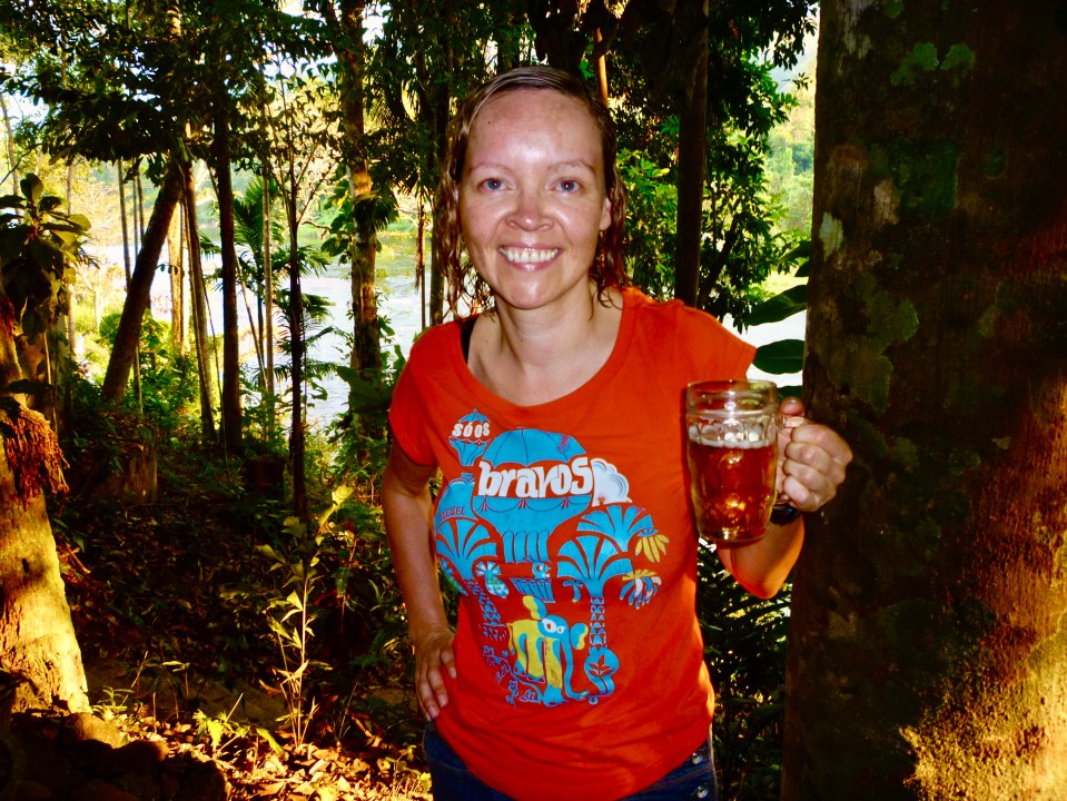 Woman in orange shirt holding a glass of beer in a forest setting.