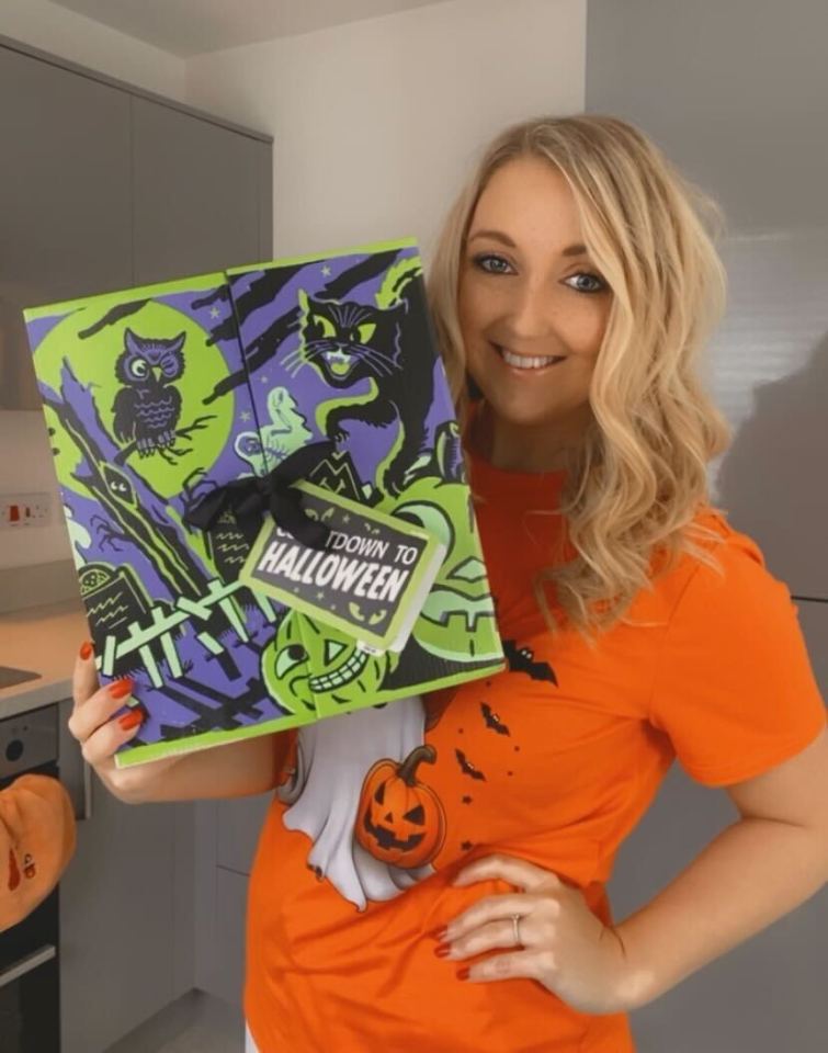 A woman in an orange Halloween t-shirt holds a green and purple Halloween box with a black cat, owl, and pumpkins.