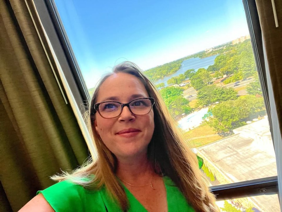 Carrie Henn in a green top looking at the camera with a lake and trees outside a window behind her.