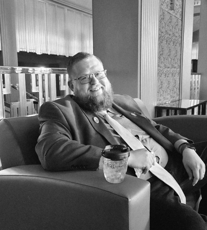 Logan Gifford, a man with a beard and glasses, smiles while seated on a couch.