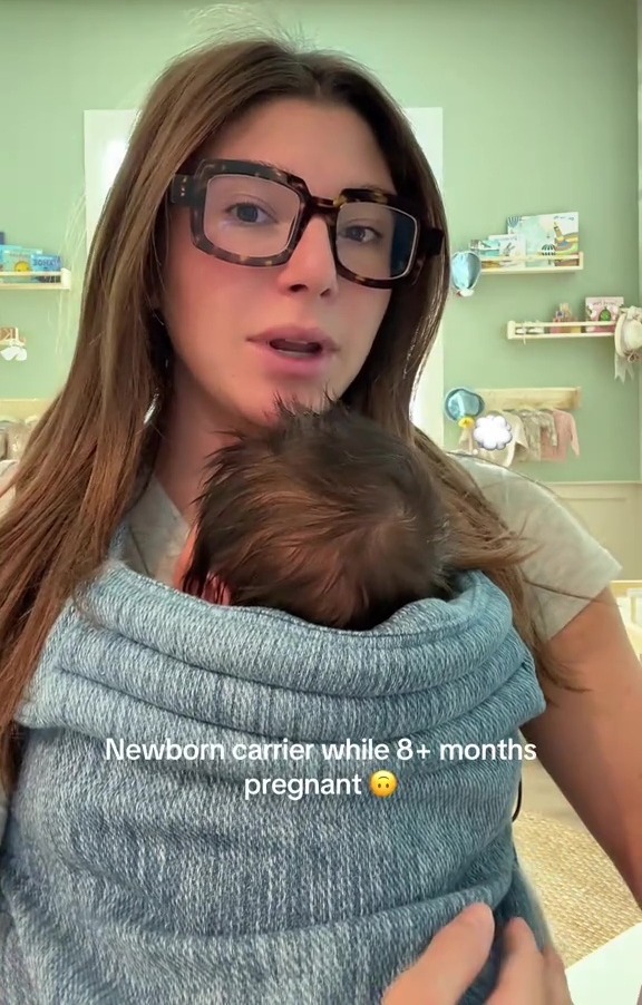 A woman wearing glasses and holding a newborn in a baby carrier on her chest, with text overlay "Newborn carrier while 8+ months pregnant".
