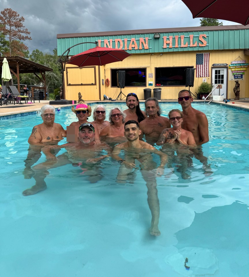 A group of 11 nude adults in a pool with a building labeled "INDIAN HILLS" in the background.
