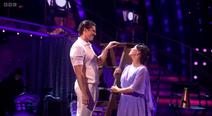 A man in a white outfit and a woman in a purple dress looking at each other, holding a wooden step stool.
