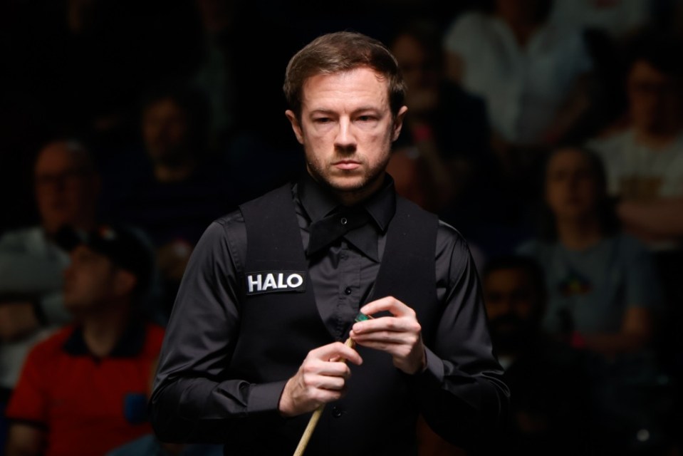 A snooker player in a black shirt and vest, holding a cue stick, looks seriously at the viewer.