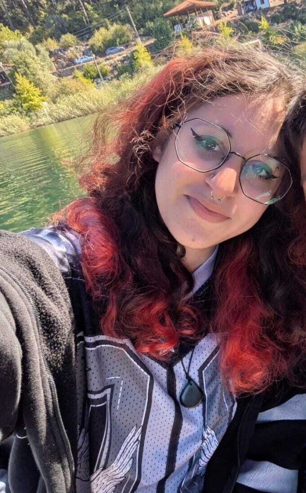 A young woman with red-streaked hair, glasses, and a septum piercing smiles in a selfie with a lake and lush hills in the background.