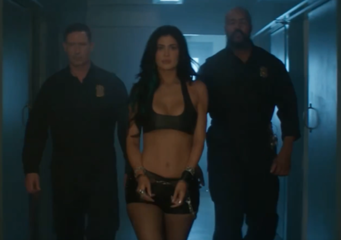 Kylie Jenner wearing a black crop top and shorts, shackled, walking with two men in dark uniforms.