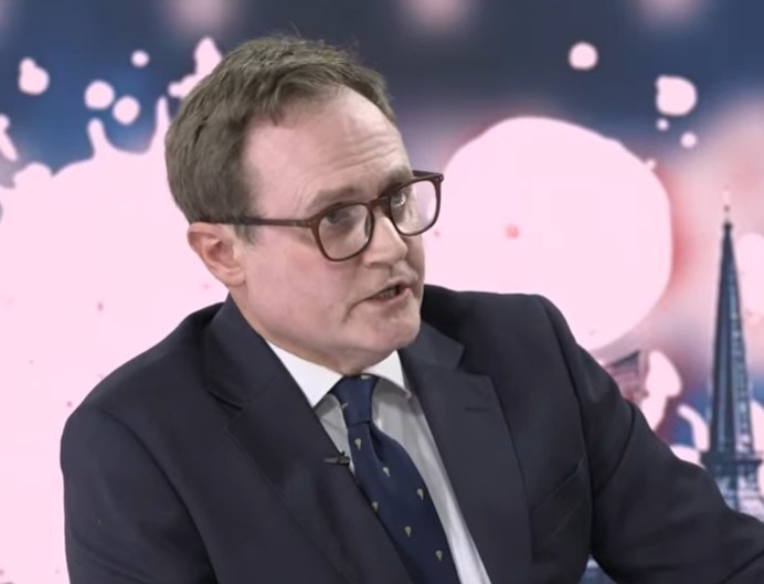 A man in a suit and tie speaks in front of a blue and red backdrop with a city skyline silhouette.
