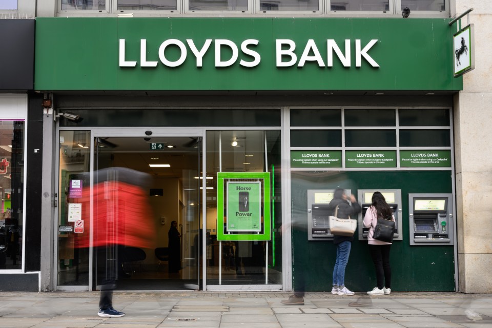 Exterior of a Lloyds Bank branch with two people using ATMs and motion-blurred figures passing by.