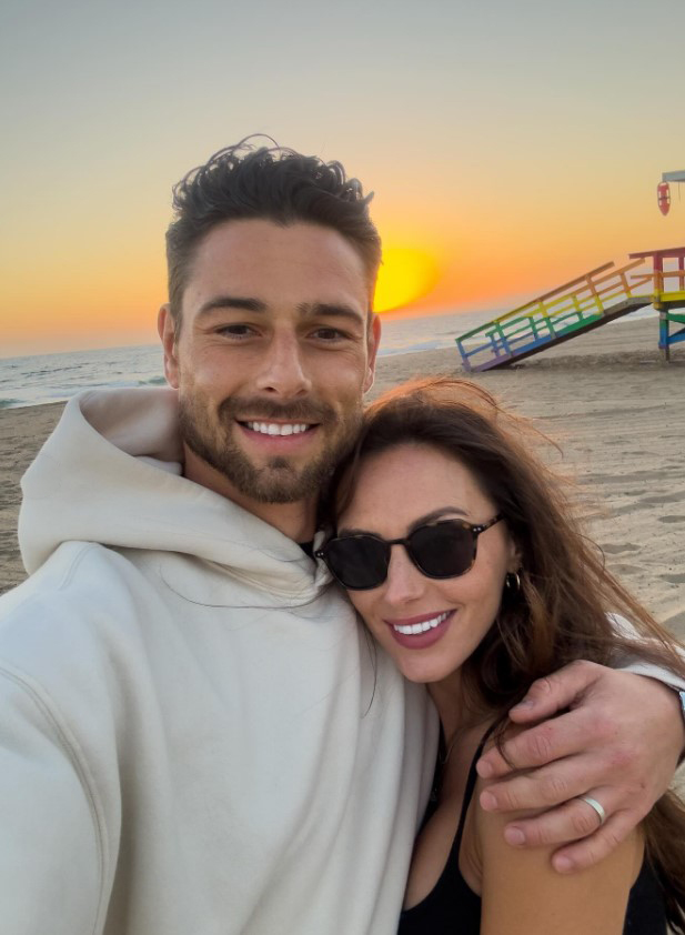 Tom Walker and Choe Sian on a beach at sunset, with a rainbow-colored lifeguard stand in the background.
