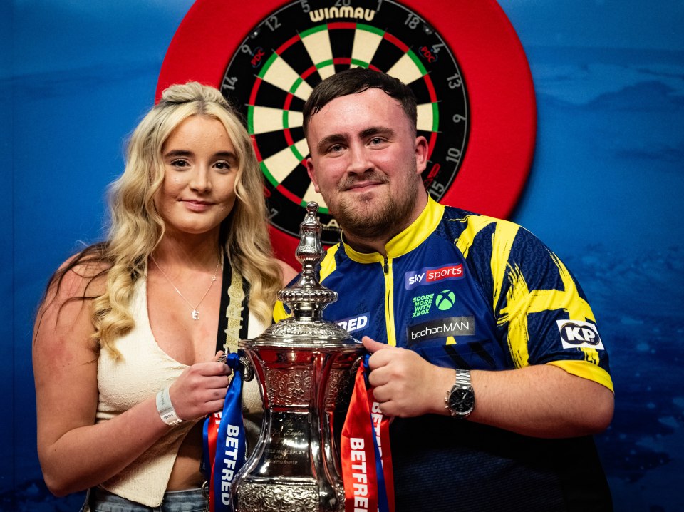 Luke Littler and his girlfriend Faith Millar holding a trophy in front of a dartboard.
