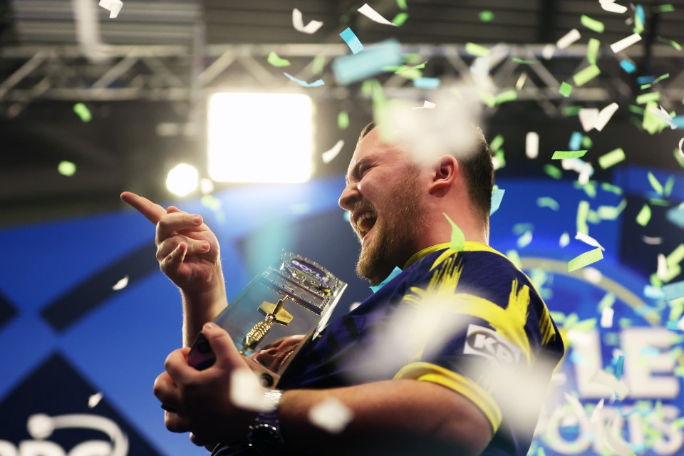 Luke Littler, a male dart player, with a beard and short dark hair, wearing a purple and yellow jersey, raises his right arm and points his finger upwards.