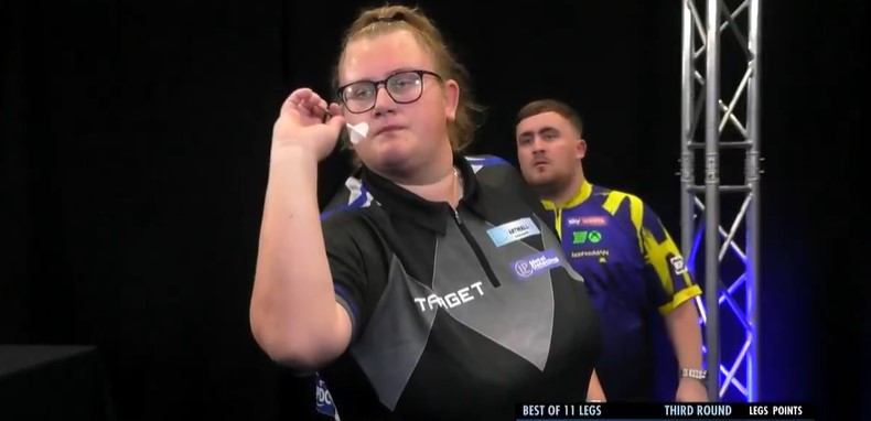 Beau Greaves about to throw a dart with Luke Littler watching behind her.