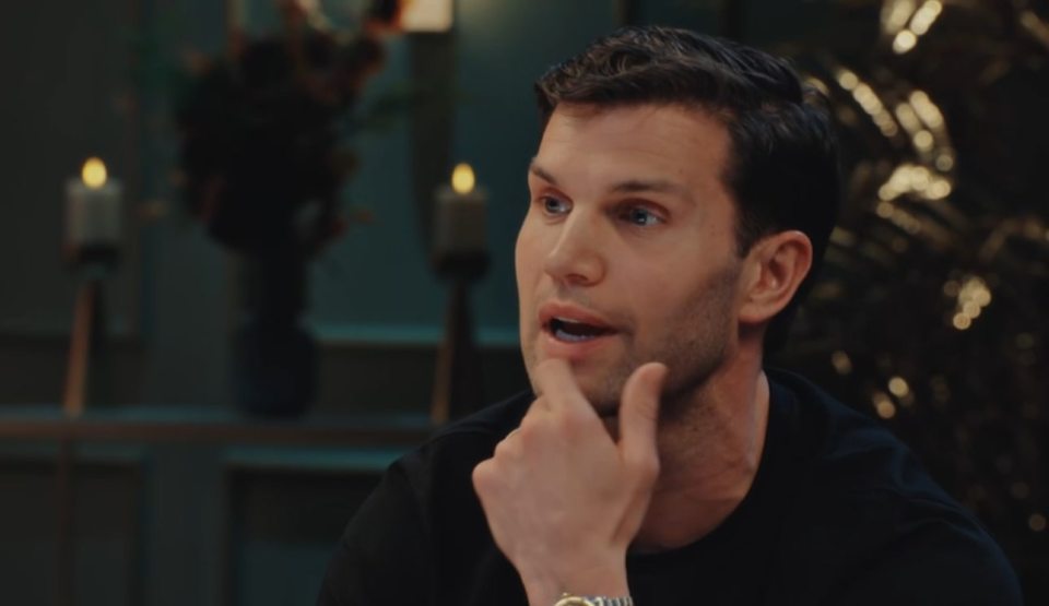 A man in a black t-shirt touches his chin while speaking.