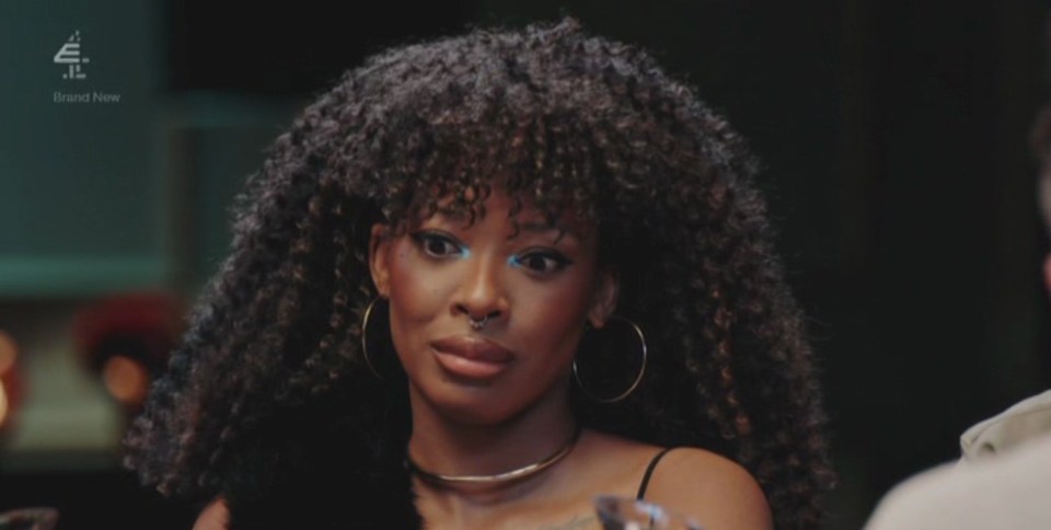 A woman with dark curly hair, hoop earrings, and a nose ring, looks to her right with a slightly confused or displeased expression.