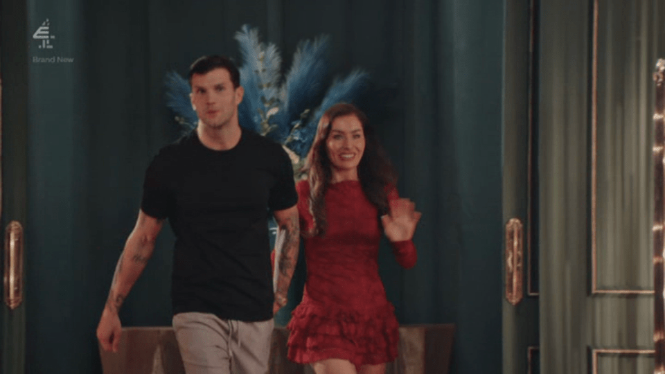 A man in a black t-shirt and grey shorts walks next to a woman in a red dress who waves.