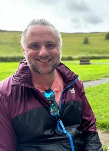 Man in a burgundy and black jacket with sunglasses hanging on the zipper, smiling in front of a green hill.